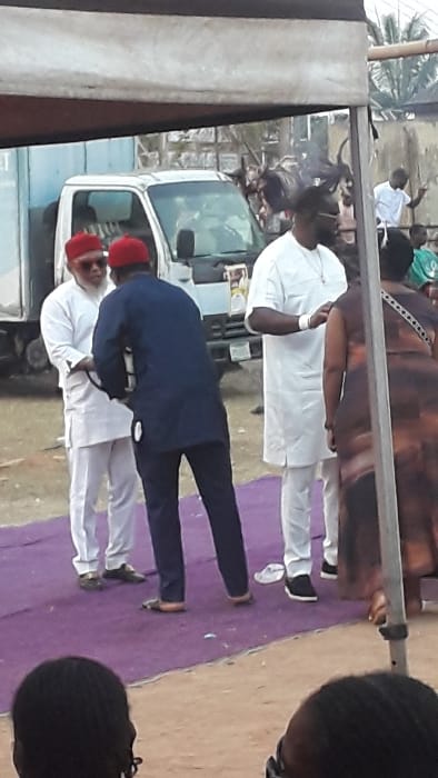 This image captures an outdoor gathering in Agukwu Nri, Nigeria. The scene is observed from a slightly low vantage point, looking towards a group of individuals under a canopy structure.
In the mid-ground, a purple carpet is laid on reddish-brown earth. On the left, two men stand facing each other. The man on the left wears a white traditional tunic and trousers with a red cap and sunglasses, holding his left hand with his right. The man facing him wears a dark blue traditional tunic and trousers with a red cap, his back partially to the camera, appearing to adjust something on the white-clad man's attire near his waist.
To the right of these two, another man in a white traditional tunic and trousers stands with dark sunglasses and a visible necklace, looking towards the right. A partially visible woman in a patterned brown dress stands to his right, her back mostly to the camera.
In the background, a white box truck with a blue rear section is parked to the left. Further right in the background, a structure with an animal (possibly a goat with horns) is visible being roasted over an open fire, producing smoke. A young male in a white t-shirt stands near this roasting setup. A light-colored concrete wall and palm fronds are also visible in the distant background. The foreground shows the heads of several unidentifiable individuals, suggesting they are seated, facing away from the camera. The scene is illuminated by natural daylight.