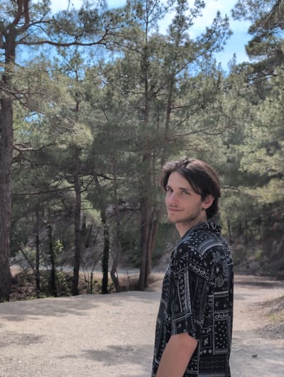 A young man with shoulder-length brown hair and a light beard, wearing a black short-sleeved shirt with a white paisley pattern, is standing in a wooded area and looking over his left shoulder towards the camera. He has blue-green eyes and is smiling slightly. The background consists of pine trees and a dirt path. The scene appears to be a natural environment, possibly a park or hiking trail, consistent with a location in Dzhankhot, Russia, a resort town known for its natural beauty and pine forests. The lighting suggests it is daytime with dappled sunlight filtering through the trees.