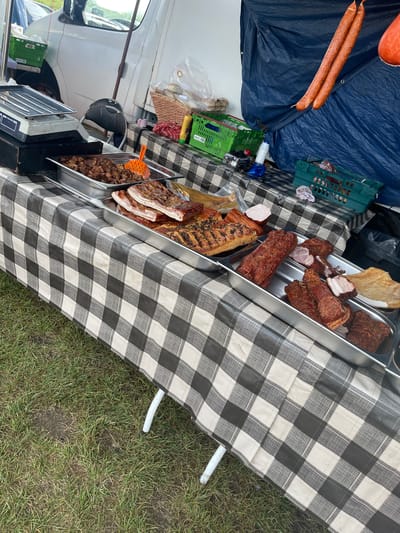 The image shows a market stall set up outdoors, likely in Sevenoaks, United Kingdom. The stall is covered with a black and white checkered tablecloth, displaying various meat products in silver trays. These include sausages, bacon, and what appears to be roasted pork belly. Behind the trays are crates, bottles, and packaging materials. A white van is visible in the background, indicating the stall is mobile. The stall's legs rest on grass.