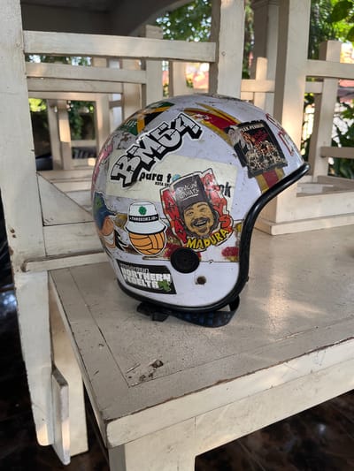 A well-worn, white motorcycle helmet adorned with numerous colorful stickers is placed on a rustic, weathered white wooden table. The stickers display various designs, including text in different fonts and languages, cartoonish illustrations, and what appear to be band logos or event flyers. One prominent sticker features a portrait of a smiling man with a mustache, wearing a distinctive hat and text in what seems to be Indonesian. Another sticker shows a green shamrock emblem with the text "THREEORZ.ID" and a stylized image of a bucket hat with a basketball pattern. The helmet is positioned at an angle, with its black chin strap visible beneath it. The table itself shows signs of age and use, with chipped paint and visible grain. In the background, the scene appears to be outdoors, with dappled sunlight filtering through greenery, suggesting a location with natural light and possibly a garden or patio area. The overall impression is of a casual, personal item placed within a relaxed outdoor setting, likely in Karangkebagusan, Indonesia, given the context.