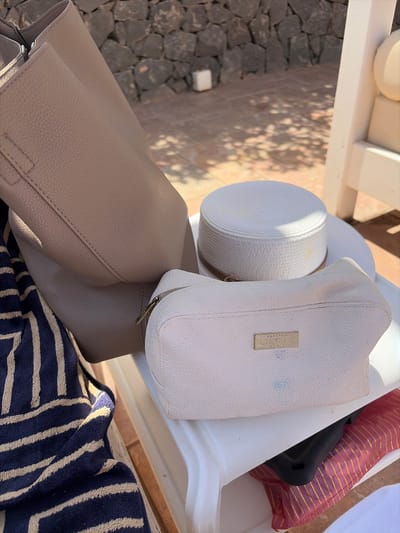 The image displays a still life of personal items on a white surface, likely a chair or table, outdoors. In the foreground, a navy blue and beige striped towel is folded, partially obscuring a large, light brown leather tote bag. Next to the tote bag is a small, light pink zippered pouch with a metallic rectangular tag that reads "LABORATOIRES LIERAC PARIS." Behind these items, a white straw hat with a brown band is positioned. The background shows a stone wall and paved ground, suggesting an outdoor setting. Sunlight casts dappled shadows on the ground and the objects. There are no people visible in the image. The scene appears to be in Antigua, Spain, as indicated by the specific location.