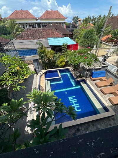 An aerial view of a swimming pool featuring the letters "WHY" written in a lighter blue shade. Lounge chairs sit beside the pool, next to some foliage and residential buildings. The buildings have red and blue roofs. Lush tropical plants are visible in the foreground and surroundings. The sky is bright blue with a few clouds, suggesting a sunny day.