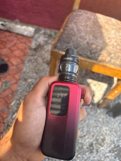 A close-up, outdoor shot captures a hand holding a red and black VOOPOO VINCI vape device, likely in Karadjovo, Bulgaria. The device features a clear tank with a visible e-liquid, and a black top section engraved with "PUSH" and "VOOPOO". The main body of the device displays "VOOPOO VINCI" on its side. Its screen is illuminated, showing various settings: "25 W", "50%", "53%", "0.30Ω", "0.8s", and "00892 P", along with the word "SMART".
The hand holding the vape is illuminated by bright sunlight, suggesting a clear, sunny day. The background is out of focus, revealing hints of an outdoor setting. Directly behind the vape, there's a blurred upholstered chair with a patterned fabric and wooden frame. Further back, the ground appears to be gravel or small stones, transitioning to a reddish paved or tiled area. The strong sunlight casts distinct highlights on the device and the hand, and potentially indicates mid-morning or afternoon.