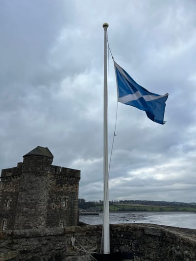 The image shows Blackness Castle in Blackness, United Kingdom, on an overcast day. A tall, white flagpole stands prominently in the mid-right, flying a blue flag with a white saltire (Scottish flag), which billows to the right. To the left of the flagpole, a portion of the grey stone castle is visible, featuring a rounded tower with a conical roof and crenellated battlements along a rectangular section. Several small, rectangular windows are set into the castle walls. In the immediate foreground, a rough, dark stone wall runs horizontally. Beyond the castle and flagpole, a wide expanse of water, likely the Firth of Forth, shows exposed muddy or sandy banks, indicating low tide. The distant shoreline features green agricultural fields, sparse trees, and a few small buildings under a uniform grey, cloudy sky. No individuals, vehicles, or animals are present.