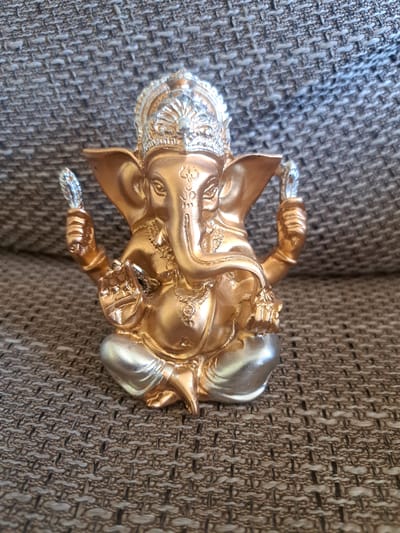 A golden and silver figurine of the Hindu deity Ganesha is positioned in the center of the frame, resting on a brown, textured fabric surface. The figurine depicts Ganesha with his characteristic elephant head, seated in a cross-legged posture. He is adorned with a crown and jewelry, and his hands are raised, holding various symbolic objects. The fabric appears to be upholstery, possibly from a sofa or chair. The background consists entirely of this textured fabric, which has a woven pattern of darker and lighter brown threads. There are no visible people, animals, or other objects in the scene. The lighting suggests a natural light source, casting subtle shadows. The location is not in Cēsis, Latvia, as the image depicts an indoor scene of a decorative object.