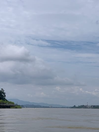 The image captures a wide, tranquil river, likely a segment of the Mekong River, with landmasses visible on either side and in the distance. The sky is heavily overcast with a mixture of light and dark clouds, creating a diffused light across the scene. On the left bank, a paved walkway or embankment is lined with palm trees and a railing, suggesting a developed area within or near the city. A few buildings and structures are discernible along this bank, blending into the distant, hazy mountains that form the backdrop. The right bank features more scattered buildings and structures, including what appears to be a taller building with a red and white striped top, possibly a communication tower or a distinctive landmark within the Golden Triangle Special Economic Zone. The river itself has a muddy, brown hue, with subtle ripples and occasional patches of lighter coloration, indicating sediment or varying water depths. A few small boats or watercraft are present on the river, suggesting ongoing activity. The overall atmosphere is calm and somewhat muted due to the cloudy weather.