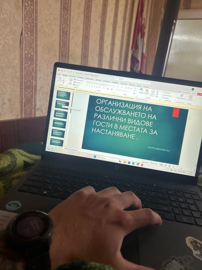 Here is a detailed description of the image:
**Scene:** The image shows a person working on a laptop, likely indoors. The screen displays a PowerPoint presentation in Bulgarian, titled "Organization of Service for Different Types of Guests in Accommodation Places."
**Objects:**
* **Laptop:** A black HP laptop is in the center, with the screen open and displaying the presentation. The keyboard is visible, and a hand is positioned over the trackpad. There are stickers on the right side of the keyboard.
* **PowerPoint Presentation:** The presentation is open in editing mode, showing the title slide and thumbnails of subsequent slides.
* **Smartwatch:** A black smartwatch is on the person's wrist.
* **Background:** The background shows a patterned wallpaper in beige and brown tones. A hint of red and white fabric is visible on the upper left edge.
* **Clothing:** The person appears to be wearing a green, textured sweater or blanket.
**People and Actions:**
* **Person:** An individual is using the laptop. The focus is on their hand, which is interacting with the trackpad.
* **Actions:** The person is presumably working on the presentation, likely editing or viewing it.
**Environment:**
* The environment appears to be a room with patterned wallpaper.
Based on the Bulgarian text in the PowerPoint, it is reasonable to assume that the image was taken in Bulgaria. There is no specific information that refers to the city of Kalchevo.