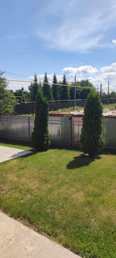 This image captures a sunny day in a suburban or rural setting, likely in Икское Устье, Russia. The scene is dominated by a well-maintained lawn in the foreground, with a concrete pathway or patio on the left. Two conical evergreen trees are planted on the lawn, casting shadows on the grass, indicating it is likely midday or early afternoon.
In the middle ground, a metal fence runs horizontally across the frame, separating the lawn from a construction or renovation area. This area appears to be partially built, with exposed bricks and debris, suggesting ongoing work. Beyond the fence, a line of taller evergreen trees forms a backdrop, with houses and more vegetation visible behind them.
The sky is a vibrant blue with scattered white clouds, typical of fair weather. Several yellow pipes, possibly gas lines, are suspended horizontally across the scene, supported by poles. These, along with the style of houses and trees, suggest a location in Russia. No people or animals are visible in the frame. The overall mood is peaceful and tranquil, conveying a sense of everyday life in a quiet residential area, possibly undergoing development. No visible text is present in the image.