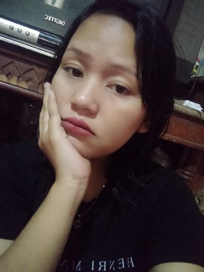 A woman with dark hair and a black shirt rests her face in her hand, looking slightly to the side. She has a serious expression. A necklace with a clover charm is visible. Behind her is a DICTLEC television, with a wooden cabinet beneath it. The lighting is soft, highlighting her skin tone and the surrounding room's details.