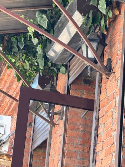 The image shows an exterior view of a building with a brick wall. Ivy plants are growing on the wall and over a metal awning structure. A dark-colored lantern with glass panels is mounted on the brick wall, beneath the awning. A metal frame, possibly for a window or door, is visible in the foreground. In the upper left portion of the image, a white rectangular object, possibly an air conditioning unit, is attached to the building. The scene appears to be outdoors, with natural light illuminating the brickwork and greenery. The presence of an air conditioning unit suggests this is likely a residential or commercial building.