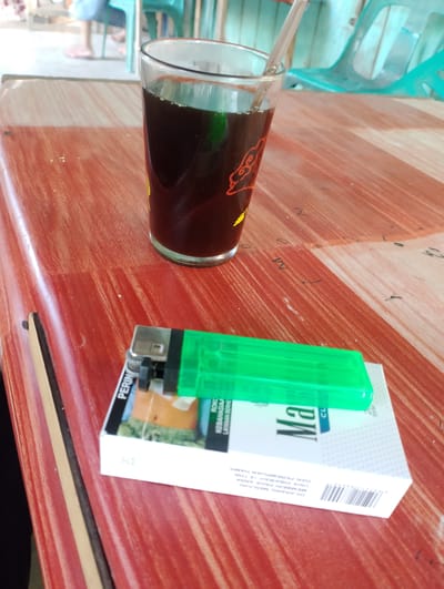 In Blangtuee, Indonesia, a close-up, eye-level shot captures a rustic wooden table. The table's surface displays a reddish-brown hue with visible wood grain and scattered dark markings. Positioned on the table are a clear glass filled with a dark beverage, likely coffee or tea, and a pack of Marlboro cigarettes. A bright green lighter rests atop the cigarette pack. The background is out of focus, showing glimpses of turquoise plastic chairs and indistinct figures of people, suggesting an outdoor or semi-open environment, possibly a warung or a casual gathering spot. The lighting is natural, contributing to a slightly grainy texture of the image.