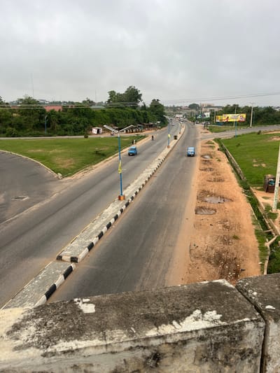 A ground-level, long shot shows a two-lane road in Osogbo, Nigeria, under an overcast sky. The road stretches toward the horizon, with vehicles visible in the distance. Roadside barriers marked with alternating black and white stripes run along the road's edge. To the left of the road, there is a grassy area with trees and buildings. To the right of the road, the terrain is sandy and uneven, with some exposed soil. In the background, billboards and other structures are visible, with power lines overhead. The foreground features a concrete structure from where the shot was taken.