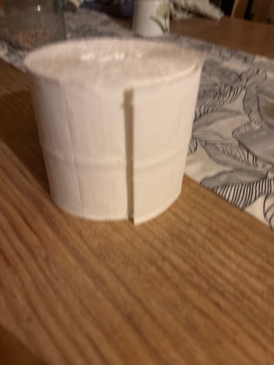 The image shows a roll of white toilet paper on a wooden table. The toilet paper is partially unrolled, with a segment of the paper peeled back. A patterned fabric, possibly a tablecloth, is visible beneath the toilet paper, and to the left, a glass jar containing a light-colored granular substance is partially visible. The scene appears to be indoors, possibly in a domestic setting. The city of Benissa, Spain, is not depicted in this image.