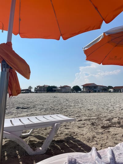 An outdoor, eye-level, medium shot captures a sandy beach scene in Skala Avdiron, Greece, under a bright, blue sky with scattered clouds. Two orange beach umbrellas are visible, one in the foreground on the left, and another in the middle ground on the right, angled slightly downwards. In the center foreground, a white plastic beach lounger is partially visible, casting shadows on the sand. The background features a row of houses and palm trees along the coastline. The sand is textured with footprints and small pebbles. The image is taken from a low angle, looking out towards the sea.