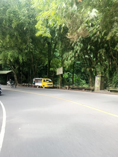 The image depicts a road in Khe Luang, Laos, lined with lush green trees and vegetation. A yellow minivan is parked on the side of the road. A motorcycle is visible on the road. The road is paved with a yellow line separating the lanes. Two people are standing near the minivan. A sign is visible next to the minivan. There's also a concrete structure. The overall environment suggests a rural setting within Khe Luang.