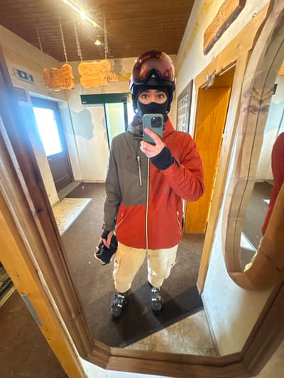 The image displays a full-body mirror selfie of an individual in an indoor hallway located in Bad Hofgastein, Austria. The person is dressed in ski equipment, wearing a two-tone red and grey ski jacket, cream-colored ski pants, and black ski boots. A black balaclava covers the lower half of their face, with a maroon ski helmet and goggles resting on top of their head. They hold a light green smartphone in their right hand, angled to capture the reflection, and a black ski pole grip with its strap in their left hand. The background, visible in the mirror, features a wooden-paneled ceiling, light-colored textured walls, and a floor composed of light-colored tiles partially covered by a dark rectangular mat. Notable environmental details include two carved wooden signs hanging from chains overhead, one partially legible as "JOLLER," a brightly lit door with a frosted glass panel on the left, a blue emergency exit sign on the left wall, and a decorative wooden door frame on the right.