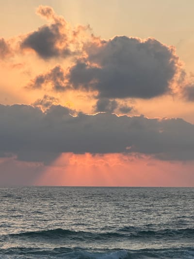 The image depicts a serene seascape at sunset, likely on the coast of Mate Asher Regional Council, Israel. The sky is a canvas of warm hues, dominated by soft oranges and pinks, with dramatic clouds casting shadows and allowing golden rays of sunlight to pierce through. The sea is a deep, textured blue, with gentle waves rolling towards the shore. The horizon is a clear, thin line separating the sky from the water, with faint silhouettes of what appear to be boats or buoys scattered across the surface. There are no people or specific structures visible in the frame, emphasizing the natural beauty of the coastal environment.