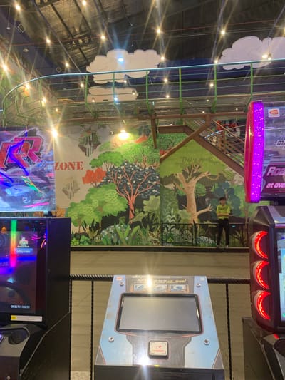 This media file captures an indoor entertainment or arcade venue in Samarinda, Indonesia, characterized by vibrant artificial lighting and themed decorations. The scene features a high ceiling with exposed structures, illuminated by numerous bright, circular lights. Cloud-shaped decorative elements are suspended from the ceiling. A prominent multi-level structure with green bars, resembling a cage or climbing frame, is visible on the left side of the image.

A large, colorful mural depicting a lush jungle landscape with various trees and foliage covers a significant portion of the wall. The word "ZONE" is integrated vertically into the mural, written in a bold, orange font. One person, wearing a light-colored shirt and dark pants, is standing casually in the mid-ground, positioned in front of this mural.

Several arcade machines are scattered throughout the venue, emitting bright, colorful lights, including purple and red glows. Text visible on these machines includes "BANDAI NAMCO," a partial game title "Roa at ove," and "up parts All...free!" Another arcade machine displays "Qualifying is in session" and "CREDIT(S)00/01." The overall atmosphere suggests a lively and engaging environment for games and recreation.