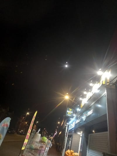 A nighttime scene captures a store front brightly lit with numerous lights stretching into the distance. A streetlamp illuminates the scene further. Shopping carts and various objects are visible near the store. In the darkened sky, a lone, distant light point shines through. The perspective is angled, adding a dynamic feel to the image.