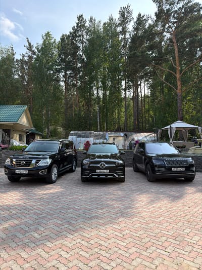 Three black luxury SUVs are parked on a brick-paved driveway in front of a building and a greenhouse. The vehicle on the left is a Nissan Patrol, identified by the "PATROL" badge on its hood and the license plate "X442XE 124". In the center is a Mercedes-Benz GLE, with license plate "C444XC 24". On the right is a Range Rover, with license plate "P710CM 124". The building appears to be a house with a green roof. Behind the vehicles, a greenhouse with a transparent roof is visible. The environment is a wooded area with tall trees. A white gazebo with draped curtains is situated in the background on the right. The sky is partly cloudy. The scene suggests a private residence in a natural setting, possibly in Sukhaya, Russia, given the license plate formatting and common SUV models found in the region.