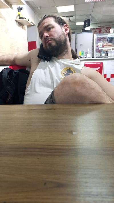 The image shows a close-up, waist-up shot of a man with long brown hair and a beard, seated at a wooden table. He is wearing a white t-shirt with a yellow and black graphic of a dragon. A black smartphone is tucked between his ear and shoulder. To his left, a black backpack is visible. The background suggests an indoor setting, possibly a fast-food restaurant, with tiled walls, a counter displaying food items, and red seating. The lighting is artificial and somewhat dim. The location appears to be within the city of Yudyino, Russia.