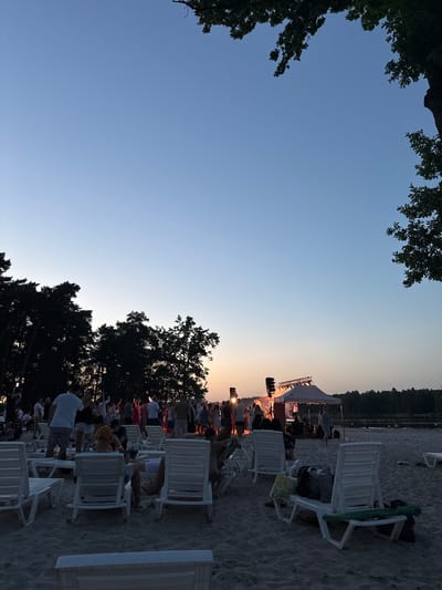 The image captures a dusky outdoor gathering on a sandy beach, likely near a body of water given the distant treeline and the presence of a lake. In the foreground, several white plastic lounge chairs are scattered on the sand, some occupied by people, while others appear empty or have bags placed on them.
In the mid-ground, a group of people are gathered, some standing and observing a stage. The stage itself is set up under a white tent-like structure and features lighting equipment and musical instruments, indicating a performance is underway or about to begin. The people are dressed in casual summer attire, and their poses suggest engagement with the event. Silhouettes of trees form a dark backdrop behind the gathering.
The sky is a clear gradient of light blue at the top, transitioning to a soft orange and pink hue near the horizon, characteristic of a sunset or twilight. The overall atmosphere suggests a relaxed, communal event. The location is identified as Знаменскі сельскі Савет, Belarus, implying a rural or semi-rural setting within the country.