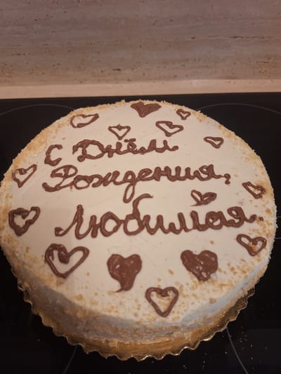 A chocolate-decorated white cake sits on a black surface. The cake is round with a white frosting and a golden, scalloped cake board. Brown icing forms the phrase "Dziękuję Kocham Cię" in elegant script, meaning "Thank you, I love you" in Polish, across the top of the cake. Small brown hearts are scattered around the text. The edges of the cake are coated with a fine crumb topping. The overall impression is a sweet and heartfelt gesture.