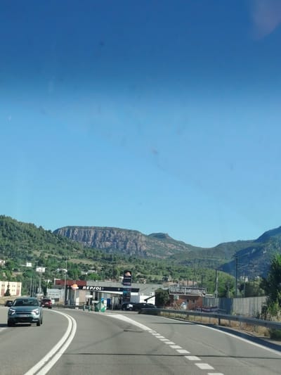 The image shows a view from a moving vehicle on a highway near Olesa de Montserrat, Spain. In the foreground, a gray car with license plate B569 LTW is driving on a highway with white lane markings. To the right of the highway is a barrier and a sign that reads "BAR RESTAURANT L'ESTRADA." Further back, a Repsol gas station with its distinctive orange and red logo is visible. Behind the gas station, a row of buildings and a mountain range covered in trees are seen. The sky is clear and blue. The overall scene depicts a typical roadside landscape with a gas station and restaurant, set against a backdrop of mountains.