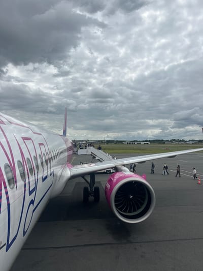 The image, viewed upright, depicts an airport scene from an airplane window, focusing on the aircraft's wing and engine against an overcast sky. The main subject is a Wizz Air airplane, easily identifiable by its distinct pink and purple livery. The word "WIZZ" is prominently displayed in large purple letters along the white fuselage, and "air.com" is visible on the pink engine cowling, which also features the Wizz Air logo.

The sky is dominated by thick, grey clouds, indicating cloudy weather. On the tarmac below, several people are seen walking, suggesting passengers are either boarding or disembarking the aircraft. A set of mobile stairs is connected to the airplane's door, facilitating access. An orange cone is also visible on the tarmac near the wing. In the background, beyond the paved ground, there is a stretch of green land and some distant structures, characteristic of an airport environment. The scene captures a moment of activity on the ground at an airport, likely Beauvais–Tillé Airport in Tillé, France, during daytime.
