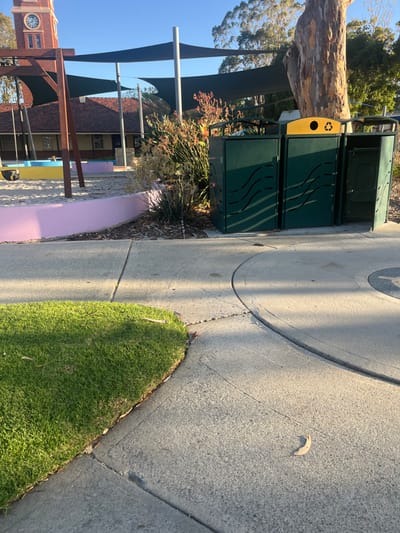 The image depicts a section of Heathcote Reserve in Applecross, Western Australia, specifically an area adjacent to a playground under clear skies.
In the foreground, a curved patch of green grass meets a light grey concrete pathway. The pathway features visible expansion joints and a single light brown leaf resting on its surface near the lower right quadrant. Diagonal shadows, likely from unseen overhead structures or trees, stretch across the concrete.
The midground features three dark green public waste bins arranged in a row along the pathway. Each bin has horizontal, wave-like cutouts on its side panels. The central bin is distinguished by a yellow top section marked with a black recycling symbol. The front panel of the rightmost bin is ajar, revealing a lighter green internal bin. Behind these receptacles, a dense cluster of green and reddish-orange shrubbery, including spiky-leaved plants, grows alongside the path. To the left of the foliage, a pink-painted concrete retaining wall outlines the edge of a sand-filled playground area.
In the background, a partial view of a wooden playground structure with a dark swing seat is visible on the far left. A large tree with light-colored bark stands prominently behind the waste bins. Three dark grey fabric shade sails, supported by tall grey poles, are positioned over the playground. Further in the background, a multi-story red-brick building with a brown tiled roof is present, featuring a distinct clock tower with a circular clock face. The sky above is clear and light blue, with numerous green trees visible on the distant horizon. No individuals are present, and no actions or interactions are depicted.