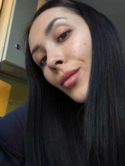 A close-up selfie of a fair-skinned young woman with long, dark hair. She has brown eyes, thick eyebrows, and is wearing lip color. The background is out of focus, but shows a neutral-colored surface with a metal button or knob, as well as a dark blue fabric. There is no indication of the location being in Molodezhnyi, Russia.