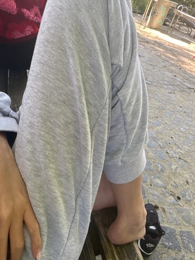 The image shows a person's legs and feet from the knee down, sitting on a wooden bench. The person is wearing grey sweatpants and a black Nike shoe on their right foot, while the left foot is bare. The background appears to be an outdoor area, possibly a park or playground, with a paved ground and some playground equipment visible in the distance. The lighting suggests it is daytime. No specific landmarks indicating Augsburg, Germany, are visible.
