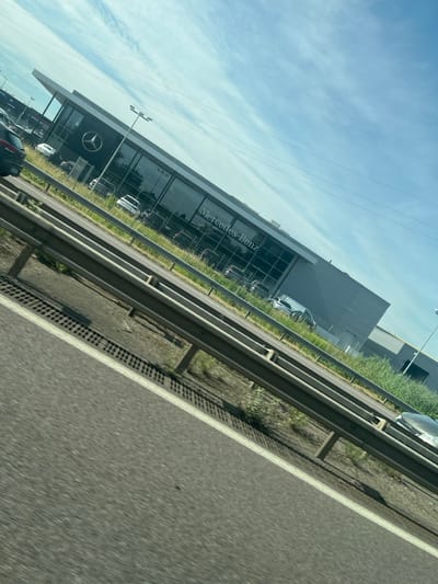 A tilted roadside view presents a Mercedes-Benz dealership with its logo prominently displayed on the building's facade. Several vehicles are visible inside the dealership. A highway with a guardrail runs adjacent to the building. The sky is partly cloudy. The photo appears to be taken from a moving vehicle.