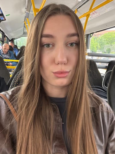 The image shows a young woman with long, straight, light brown hair taking a selfie on a bus. She is wearing a dark gray t-shirt and a brown leather jacket, with a thin silver necklace visible. Her skin is fair and she has light green eyes and light pink lips. The bus interior is visible behind her, with yellow support poles and dark gray patterned seats. Two men are seated in the background on the left side of the bus. One man is bald and wearing a blue and white checkered shirt under a blue jacket, and the other man is wearing a blue shirt. Outside the window, trees and buildings are visible, suggesting an urban or suburban setting. The lighting suggests it is daytime. The image appears to be taken within the city of Sissach, Switzerland.