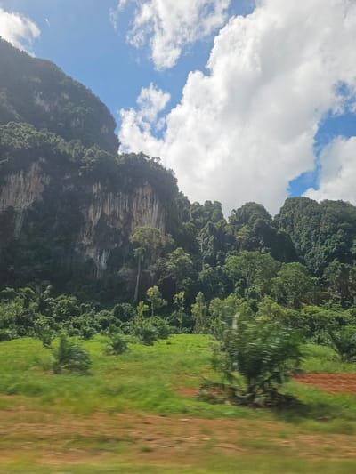 The image depicts a dramatic natural landscape, likely taken from a moving vehicle, as evidenced by the motion blur on the right side of the frame. The scene is dominated by a steep, verdant mountain covered in dense tropical foliage. The mountain's sheer rock face, characterized by layered striations, is partially visible, suggesting a geological formation of significant age. Above the mountain, a bright blue sky is dotted with fluffy white clouds, indicating fair weather.

The foreground on the right is a blur of green and brown, likely representing a field or plantation adjacent to the road. The overall impression is one of lush, untamed natural beauty, possibly in a region with a tropical climate, consistent with the provided location context of Ban Nai Sa, Thailand. The time of day appears to be daytime, given the brightness of the sky and the illumination of the landscape. There is no text visible in the image. The scene evokes a sense of awe and the grandeur of nature.