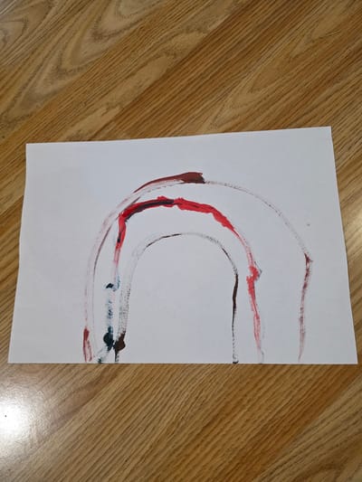 A piece of white paper with an abstract painting lies on a light brown wooden surface, likely a floor or table, in Hadzhidimitrovo, Bulgaria. The paper is oriented vertically, and the wood grain runs diagonally from the upper left to the lower right.
The painting consists of several broad, curving paint strokes, primarily forming a large, irregular 'C' or horseshoe shape that extends from the lower-left to the middle-right of the paper. The dominant colors used are various shades of red and a dark reddish-brown, almost maroon. Within the main curve, a vibrant red stroke stands out as the thickest and most prominent. Other, thinner lines of red and brown run alongside it, some appearing layered. Towards the right side of the painting, streaks of dark blue or teal paint are visible, mixing with the red and brown tones. The paint strokes show varying thickness and texture, indicating the use of a brush and suggesting different amounts of paint applied. There is a small, faint grey smudge near the top-center of the paper.
The lighting is bright and even, suggesting an indoor setting during the daytime or a well-lit interior. No discernible text is visible on the paper or in the background.