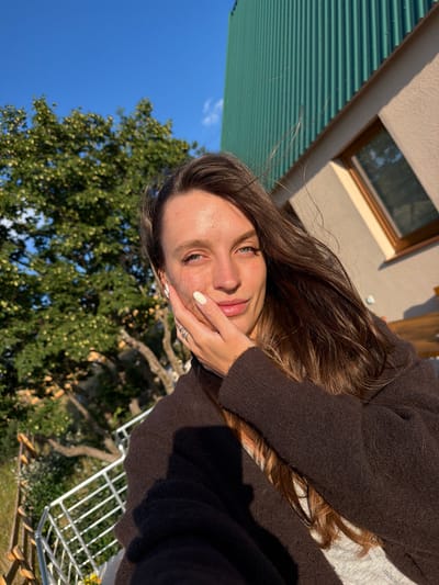 A young woman with long, dark brown hair and blue eyes is captured in a close-up, outdoor selfie. She is positioned on a balcony with a railing in the foreground, and a wooden fence behind it. To her left, a lush green tree with scattered yellow blossoms is visible against a bright blue sky. The background to her right features a building with a corrugated green roof and a light-colored wall with a wooden-framed window. The woman is wearing a dark brown cardigan over a light-colored top. Her right hand is gently touching her cheek, and she is wearing a silver ring on her left ring finger and has white-painted fingernails. The lighting suggests it is late afternoon or early evening, with the sun casting a warm glow on her face and the surroundings. The location is identified as Bukovac, Montenegro.