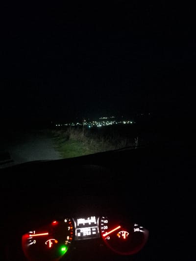The scene is a nighttime view from inside a stationary vehicle located on an unpaved path overlooking Krumovo Gradishte, Bulgaria. In the foreground, the vehicle's illuminated dashboard displays a tachometer at approximately 0 RPM, a speedometer indicating 0 km/h, a digital readout of "271.0," and an active low fuel warning light. A green indicator light is also illuminated. The vehicle's headlights project light onto the immediate unpaved path and adjacent grassy terrain. The distant background features a cluster of artificial lights, characteristic of a town, representing Krumovo Gradishte, under a dark sky. No individuals are visible.