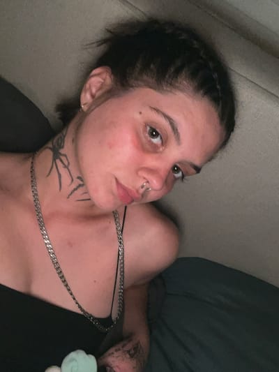 A young person with dark hair styled in a braid is lying down, facing the camera at an angle. They have a spider tattoo on their neck and a silver chain necklace. They are wearing a black tank top. The background appears to be a bed with a textured headboard. The lighting is dim, suggesting an indoor setting, possibly a bedroom. There is no indication of the city of Genemuiden, Netherlands.