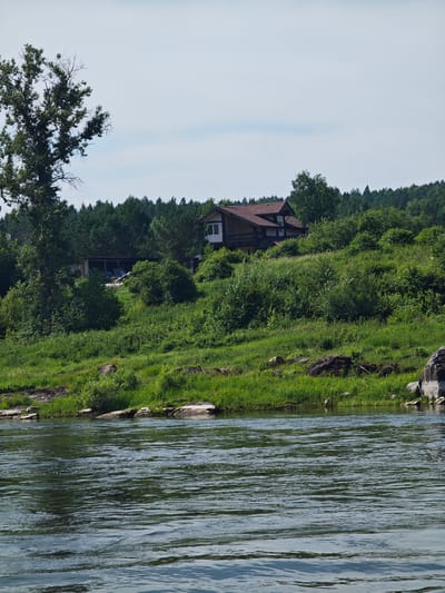 The image depicts a serene landscape featuring a wide river in the foreground, its surface rippling under a clear sky. The riverbank is lined with lush green grass and interspersed with large rocks. In the mid-ground, a dense forest of evergreen trees forms a backdrop. Nestled on a gently sloping hill amidst the foliage is a modern, multi-story house with a dark, pitched roof and light-colored walls, featuring multiple windows. To the left of the house, a dilapidated structure with a corrugated roof and open sides stands as a stark contrast to the well-maintained residence. A tall, leafy tree with a bare upper section stands prominently to the left of the house. The scene is likely located in or near the city of Porog, Russia, given the overall natural environment and architectural style.