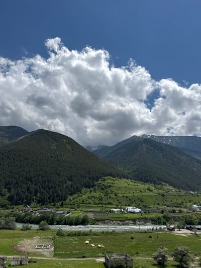 This is a landscape photograph taken in Верхняя Теберда, Russia. The image captures a wide view of a valley with mountains, a river, and a village. In the foreground, there is a grassy field with some partially constructed or ruined buildings and stacks of lumber. A dirt road runs through the field.
A wide, turquoise river flows through the midground, bordered by trees and a grassy bank. On the other side of the river, a village is nestled, characterized by various houses with dark roofs, some larger buildings, and fenced areas. The village is situated at the base of rolling green hills, which transition into steeper, forested mountains.
The mountains dominate the background. The closest mountain on the left is densely covered with dark green coniferous trees, its slopes rising sharply. Further back, other mountain ranges are visible, with some showing patches of snow on their upper reaches, indicating a higher altitude. The sky above is a bright blue, with large, fluffy white clouds scattered across it. The lighting suggests it is daytime, likely late morning or early afternoon, with the sun illuminating the landscape. The overall scene depicts a serene, rural mountain environment.