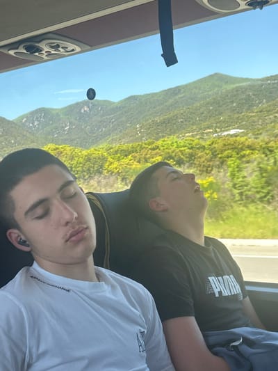 Two men are seated on a bus with a landscape view of Vrasna, Greece, seen through the window. One man, wearing a white Armani Exchange shirt, is resting with his eyes closed. The other man, wearing a black Puma shirt, is also resting with his eyes closed. A dashboard is visible with a control panel and a black strap extending outward. An emblem is affixed to the window. The background shows a mountainous, green landscape under a blue sky.