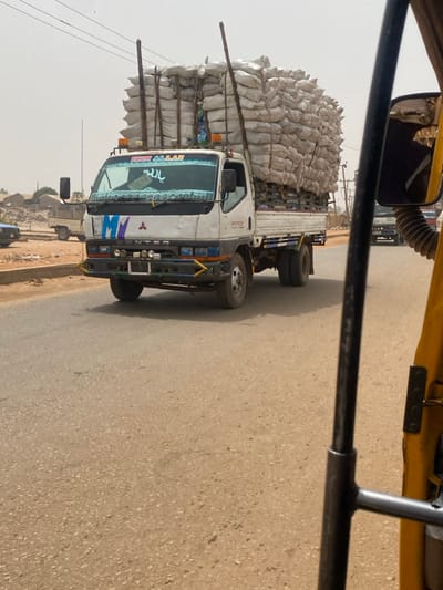 A white Mitsubishi Fuso Canter truck, extensively overloaded with numerous light-colored sacks stacked multiple layers high, is depicted driving on a paved road in the city of 0801712591, Nigeria. The cargo is secured by vertical wooden poles extending above the truck's cabin. The vehicle's windshield features Arabic script 'يا الله' and the text 'IRON ALLAH' on a visor. The front bumper is a customized assembly, painted blue and yellow, equipped with auxiliary lights. The image is captured from the interior perspective of a yellow vehicle, likely a tricycle, with its frame and side mirror visible on the right. The road surface is dry asphalt. In the background, low-rise buildings, utility poles, and distant vehicles are visible under a hazy, light-colored sky, indicating an urban or semi-urban Nigerian environment. No specific individuals are discernibly visible.