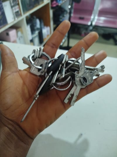 A person's hand, displaying prominent veins and skin texture, holds a bunch of keys. The keys are made of silver-colored metal and vary in design, with some featuring intricate teeth and others having simpler cuts. One key has a black plastic head, suggesting it might be a car key or a remote key. The keys are attached to a metal ring. The background is blurred but suggests an indoor setting. Shelves with various items, some appearing to be boxed products, are visible on the left. On the right, a pink object, possibly part of a vehicle, is discernible. The lighting appears to be artificial, indicating the photo was taken indoors, likely during the day. The overall scene is casual, with the focus entirely on the keys and the hand holding them. There is no visible text in the image.