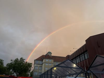 An outdoor scene captures a vibrant rainbow arching across a cloudy sky, with urban buildings and trees in the foreground. The sky transitions from a somber grey on the far left to a warm, soft ochre and light brown hue on the right, providing a striking backdrop for the rainbow's full arc. The rainbow displays a clear spectrum of colors, appearing brightest against the warmer sky.

In the mid-ground, a prominent older multi-story building features a light tan facade, multiple rectangular windows, and a distinctive reddish-brown tiled roof. A small, green-domed cupola adorns its top, and a spire rises from the roof. To its right, a more modern structure with dark red-brown cladding and dark-framed windows is partially visible. In the immediate foreground, several contemporary glass canopy structures with dark frames are seen, some showing reflections.

Lush green trees are visible on the far left, their dense foliage providing a contrast to the muted sky and buildings. On a red structure partially obscured by the trees, fragmented text is discernible: "WERGRILLHAUS" and "ÖBEL", suggesting commercial establishments like a grill house or furniture store.

The diffused light and warm tones in the sky indicate either late afternoon or early evening. The presence of the rainbow confirms recent rainfall accompanied by sunshine, creating a serene and picturesque atmosphere over the urban landscape of Landau in der Pfalz, Germany. No people are visible in the image.