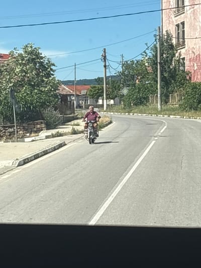 An elderly man is riding a motorcycle on a curved road in a village. The man is wearing a maroon shirt and appears to be Caucasian. The motorcycle is silver with a black seat. The road has a white line painted down the center. To the left of the road, there is a sidewalk made of stone and concrete. Behind the sidewalk, there are some stone walls and green bushes. Further back, there are houses with red tile roofs and trees. To the right of the road, there is a pink building with windows and a fence made of vertical posts. The sky is blue and clear. The overall impression is a rural setting on a sunny day. The scene is located in the village of Mladinovo, Bulgaria.