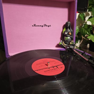 The image presents a close-up, slightly elevated view of a vinyl record playing on a portable record player in Kladovitsy, Russia. The record player's interior lid is pink and features the script text "Sunny Days," while its visible exterior is purple. A black vinyl record with a bright red label is centered on the turntable, with the tonearm and needle positioned, suggesting active playback.
To the right, a striking dark-skinned female figurine is prominently displayed. She has distinctive green and yellow highlights in her long hair, and celestial symbols—a crescent moon on her left breast and a sun on her right—adorn her chest. She cradles a form in her lap, possibly a child or an earth-like creature, also accented with green and yellow. Behind the figurine, lush green, ivy-like foliage is visible, contributing to a natural, somewhat mystic ambiance. The lighting is dim and warm, creating subtle reflections on the shiny surface of the vinyl record.
The red record label contains significant Cyrillic text. It identifies the record company as "МЕЛОДИЯ" (Melodiya), a Soviet-era label, and the manufacturer as "ЛЕНИНГРАДСКИЙ ЗАВОД" (Leningrad Plant). The artist is named "ПАРКЕР" (Parker), with "Ч. Паркер" (C. Parker) specified, strongly indicating jazz musician Charlie Parker. The label notes "Записи 1940-х, 1950-х гг." (Recordings of 1940s, 1950s) and "Выпуск 1988 г." (Release 1988), suggesting a later compilation of earlier jazz works. Other visible details include "ГОСТ 5289-80" (State Standard) and technical numbers like "M60 48437 007" and "МОНО 33".