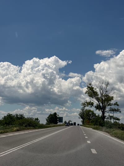 The image depicts a road near the city of Makariopolsko, Bulgaria. The sky occupies the left portion of the frame, showing a bright blue area contrasting with the large, billowing cumulus clouds. On the right, the asphalt road stretches into the distance, marked with a double white line and dashed lane markings. A line of cars is visible on the road. The roadside is bordered by green foliage and trees. A small sign, probably a traffic sign, is visible near the road.