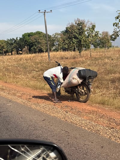 The image depicts an outdoor scene in Bokana, Nigeria, under a clear, sunny sky. A dark-skinned male, wearing a white t-shirt with red and yellow accents and dark blue sweatpants with yellow stripes, is bent over a stationary silver motorcycle positioned on a dirt shoulder beside an asphalt road. He is barefoot, with black sandals on the ground near his feet, and appears to be interacting with the motorcycle's engine or frame area. The motorcycle is heavily laden with cargo, including a large, light-colored sack secured to its rear and a black elongated object resting on top of the sack. The ground surrounding the motorcycle and stretching into the background consists of dry, golden-brown grass. Scattered trees, including some palm-like species on the left and deciduous trees further right, populate the landscape. A grey utility pole with overhead power lines stands in the mid-background. The lower left foreground reveals a portion of a fragmented car side mirror.