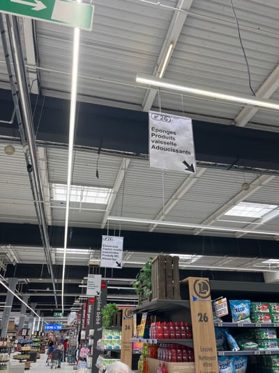 The image shows the interior of a supermarket in Saint-Brice-Courcelles, France. The location is marked by the coordinates: latitude 49.283, longitude 3.999. The image focuses on a sign hanging from the ceiling, with the number "26" printed on it. The sign reads, "Éponges Produits vaisselle Adoucissants" (Sponges Dishwashing Products Softeners) and points to the right with a black arrow. The sign is located above aisle number 26.
There are numerous other signs hanging from the ceiling, indicating other product locations, such as aisle number 24 which features "Essuie-tout Papters toilette Incontinence Produits PVC Allume feu" (Paper towels Toilet paper Incontinence PVC products Fire starter).
There are also signs with the letter "L" in a circle, potentially the logo for the supermarket chain. The aisle in the image is filled with various cleaning products, mostly bottles of red liquid, stacked on shelves, as well as a few cleaning supplies and a sign for "Lessive Nettoyant vitres Linettes" (Laundry detergent Glass cleaner Linens). The environment appears to be brightly lit and modern, with a metal ceiling grid, metal beams, and skylights. The camera angle is pointing slightly upward towards the ceiling. It is difficult to determine if there are any people in the image, due to the low image resolution.